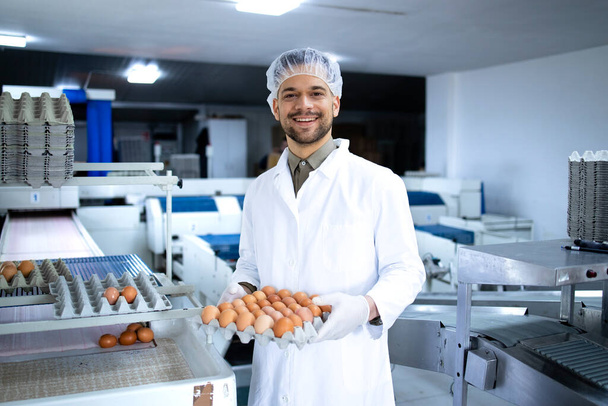 Portret pracownika farmy jajecznej trzymającego kartonową skrzynkę ze świeżymi jajami. W tło maszyny transportującej jaja do pakowania. - Zdjęcie, obraz