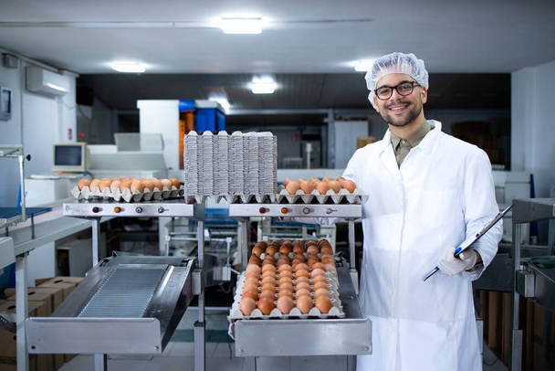 Portrait de technologue d'usine alimentaire avec filet à cheveux et gants hygiéniques debout par la machine industrielle de transport et d'emballage des oeufs dans l'usine de transformation des aliments. - Photo, image