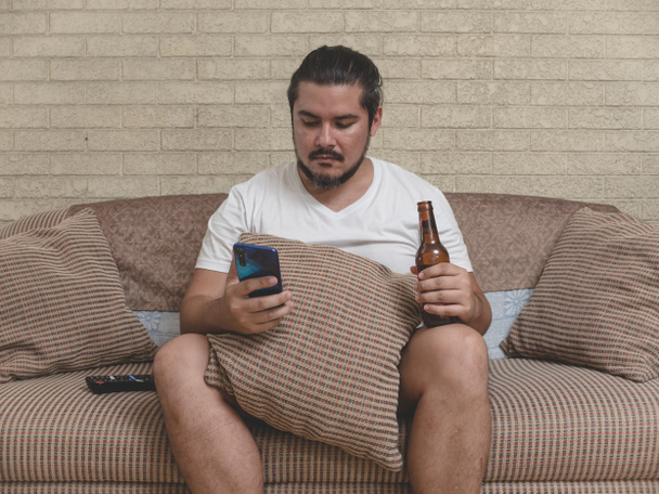 Ein fauler Mann in seinen Dreißigern blättert in seinem Handy, während er sich auf die Couch setzt und eine Flasche Bier in der Hand hält. Freizeit- oder Lebensstilkonzept. - Foto, Bild