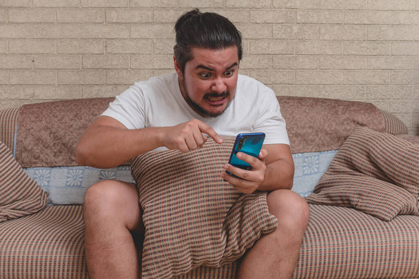 Un hombre enojado y desencadenado despotrica y regaña a alguien en el videocall en el teléfono mientras está sentado en el sofá en casa. - Foto, Imagen