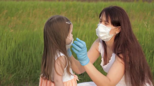 Anya maszkot és kesztyűt visel, orvosi maszkot tesz a gyerekre nyáron az utcán a koronavírus járvány idején. Anya és lánya orvosi maszkban a szabadban. Boldog család anya, lánya ölelés - Felvétel, videó