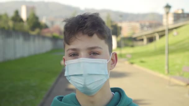 Ein ernst besorgter junger Mann in medizinischer Maske blickt in einem Park mit Vegetation und Rasen vor dem Hintergrund einer Brücke und des Verkehrs auf. Coronavirus und Allergieschutzkonzept - Filmmaterial, Video