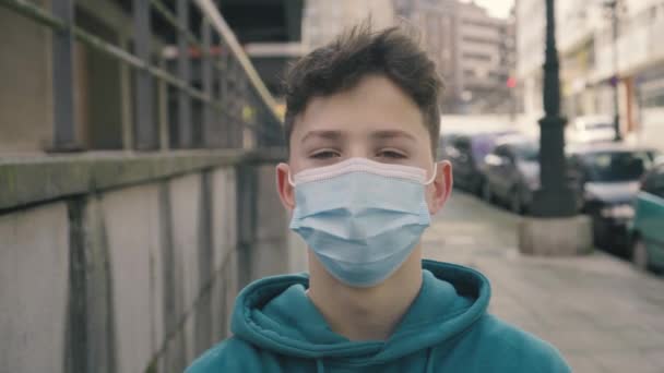 Ένας έφηβος με μια ιατρική μάσκα μιας χρήσης στο δρόμο με σταθμευμένα αυτοκίνητα. Χαμόγελα. Έννοια της ατμοσφαιρικής ρύπανσης και πρόληψη των κορωναϊών - Πλάνα, βίντεο