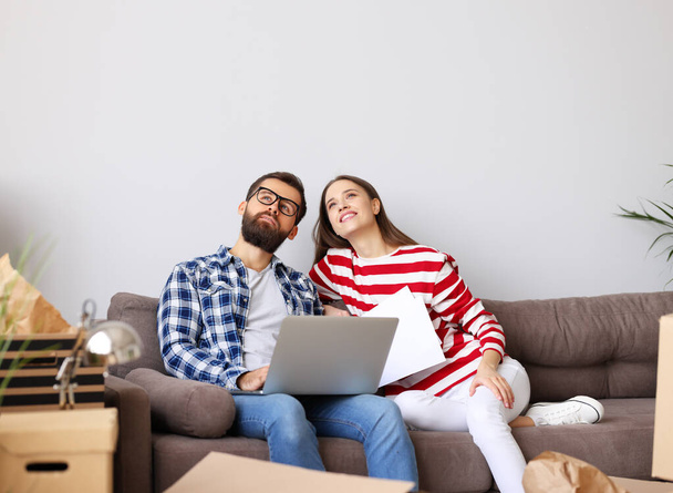 Full body of happy young man with laptop on knees  while sitting with wife   among packed stuff and discussing ideas about interior design of new home - 写真・画像