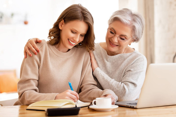 Mladá žena v neformálním oblečení dělat poznámky v plánovači, zatímco pomáhá starší matka pracuje na dálku na notebooku - Fotografie, Obrázek