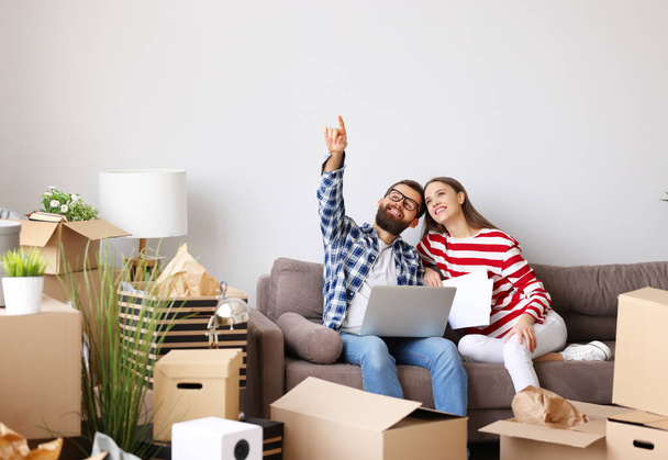 Full body of happy young man with laptop on knees  while sitting with wife  pointing up among packed stuff and discussing ideas about interior design of new home - Photo, Image
