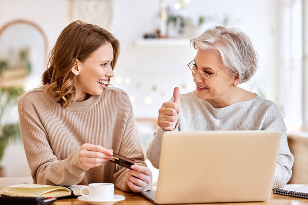 Joven hija sonriendo y mirando a anciana dama con tarjeta de crédito, mientras que las compras en línea en casa juntos - Foto, Imagen