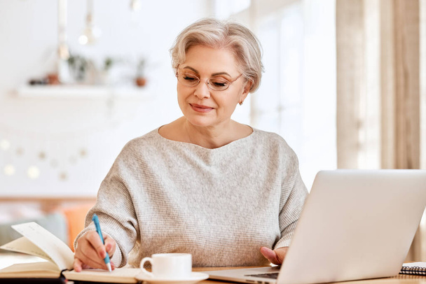 Senior vrouw met laptop en notebook terwijl zitten aan tafel en werken aan remote project thuis - Foto, afbeelding