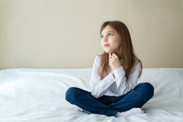 Close up shot of beautiful blonde caucasian little girl wearing jeans and a white shirt, sitting on a bed and looking calm and dreamy  - 写真・画像