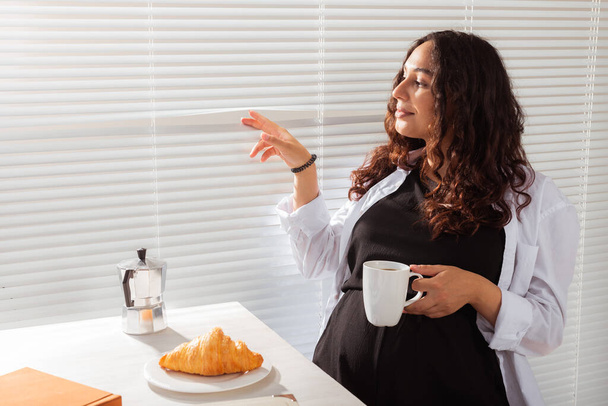 Joyful zwangere jonge mooie vrouw kijkt door jaloezieën tijdens haar ontbijt met koffie en croissants. Begrip goedemorgen en wachten op een ontmoeting met een baby. Kopieerruimte - Foto, afbeelding