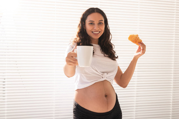 Mooie zwangere vrouw met croissant en een kop koffie in haar handen tijdens het ontbijt. Concept van goede gezondheid en positieve houding tijdens de bevalling - Foto, afbeelding