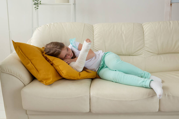 Gyermek gipsszel a törött csuklóján vagy karján a kanapén fekszik. Helyreállítás és a betegség fogalma. - Fotó, kép