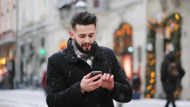 Un hombre va a la plaza central en una nevada. Él está enviando mensajes de texto en su teléfono inteligente y sonriendo. 4K - Metraje, vídeo