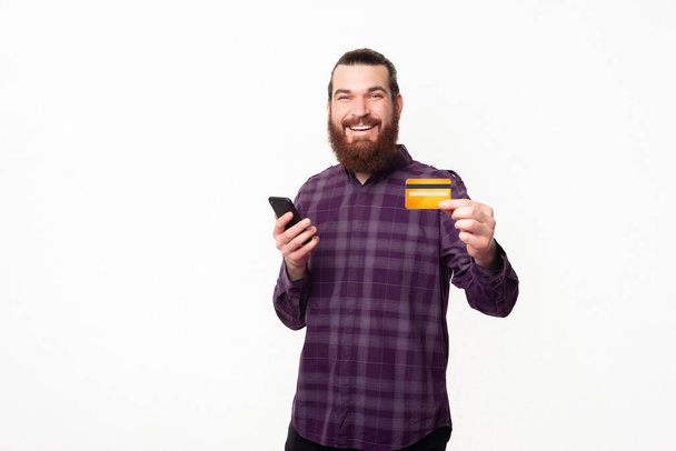 Joyeux jeune homme barbu décontracté montrant carte de crédit jaune et tenant smartphone. - Photo, image