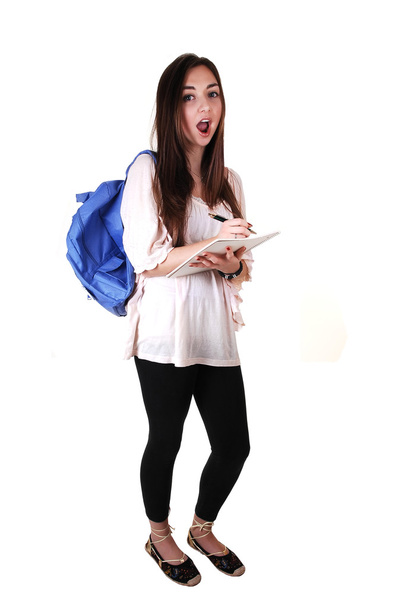 Uma adolescente surpreso com uma mochila azul sobre o ombro em uma blusa bege e calças pretas, com um caderno na mão, sobre branco
. - Foto, Imagem