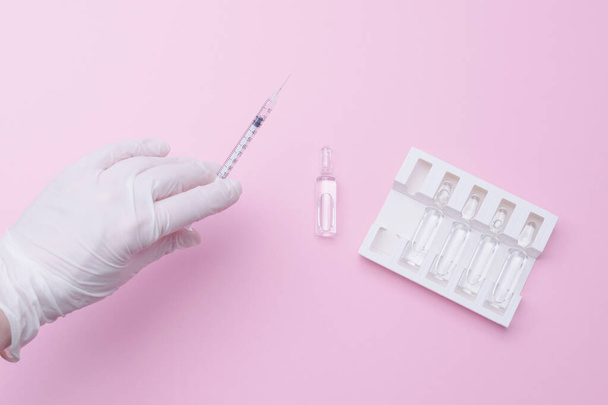 Εικόνα του χεριού ενός γιατρού σε ένα λευκό προστατευτικό ιατρικό γάντι που κρατάει μια σύριγγα με ένα φάρμακο, ένα εμβόλιο, πάνω από ένα τραπέζι με ένα σετ αμπούλες. Οριζόντια φωτογραφία, ροζ φόντο, θέση για το κείμενό σας. - Φωτογραφία, εικόνα