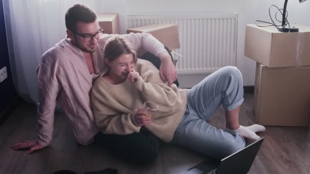 Happy couple relaxes after moving in a new home. Couple sits on floor and watches comedy movie on notebook - Footage, Video