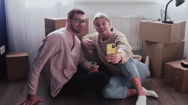 Happy couple relaxes after moving in a new home. Couple sits on floor and celebrates their moving in a new home. Drinks an alcohol and talks a selfie - Footage, Video