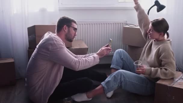 Happy couple relaxes after moving in a new home. Couple sits on floor and celebrates their moving in a new home. Man drinks an alcohol and takes a photo of his wife - Footage, Video