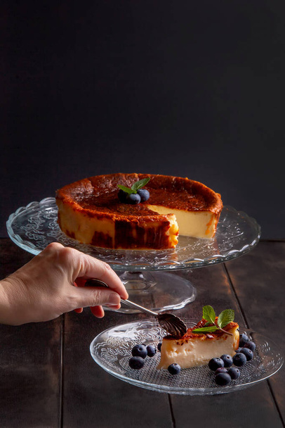 Domácí baskický zapálený tvarohový koláč se smetanovým sýrem, vejci a smetanou pečené v drobenkové kůře, s borůvkami a mátovými listy. Žena zlomí kus na talíři na tmavém pozadí. - Fotografie, Obrázek