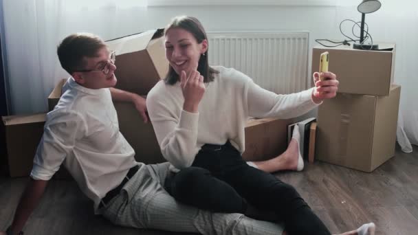 Happy couple relaxes after moving in a new home. Couple sits on floor and celebrates their moving in a new home. They records video selfie on phone - Footage, Video