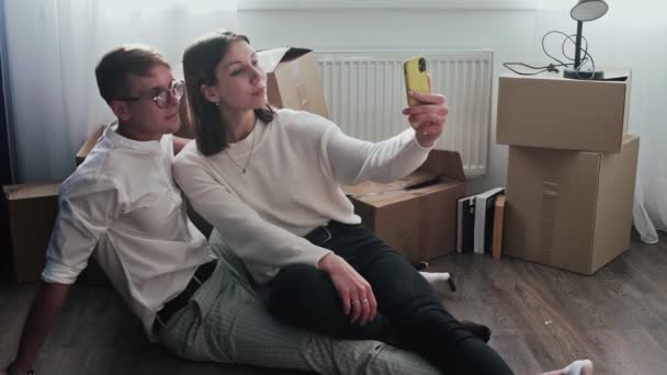 Happy couple relaxes after moving in a new home. Couple sits on floor and celebrates their moving in a new home. They takes a selfie on phone - Footage, Video