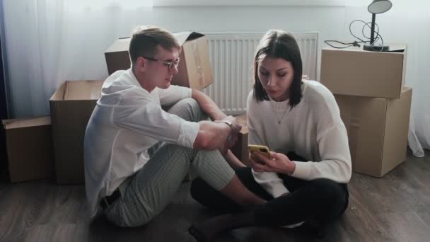 Happy couple relaxes after moving in a new home. Couple sits on floor and woman talks on phone - Footage, Video