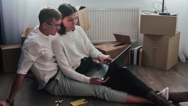 Šťastný pár se uvolní po přestěhování do nového domova. Pár sedí na podlaze a používá notebook pro výběr nového nábytku v novém domově - Záběry, video