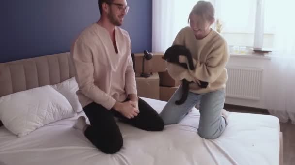 Happy couple relaxes after moving in a new home. Couple have fun on bed with a black cat - Footage, Video