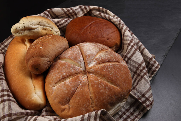 Pan recién horneado pan pan rollo redondo larga mezcla verity envuelto en tela de cocina a cuadros toalla de servilleta sobre fondo negro - Foto, imagen