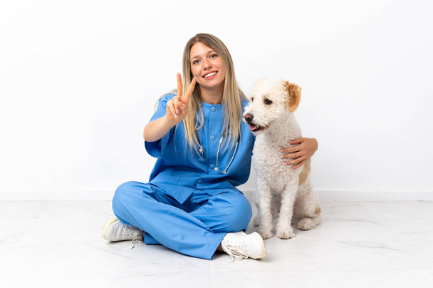 Fiatal állatorvos nő kutyával ül a padlón mosolyog és mutatja győzelem jele - Fotó, kép