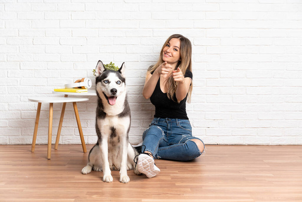 Fiatal csinos nő a husky kutya ül a padlón beltéri mutat ujjal rád egy magabiztos kifejezés - Fotó, kép