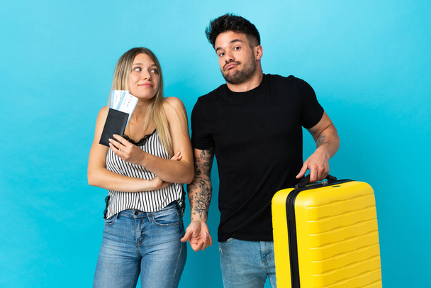 Young caucasian couple going to travel isolated on blue background making doubts gesture while lifting the shoulders - Photo, Image