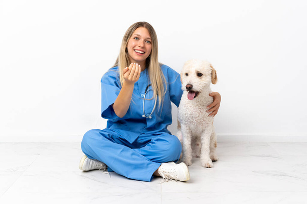 Young veterinarian woman with dog sitting on the floor inviting to come with hand. Happy that you came - Zdjęcie, obraz