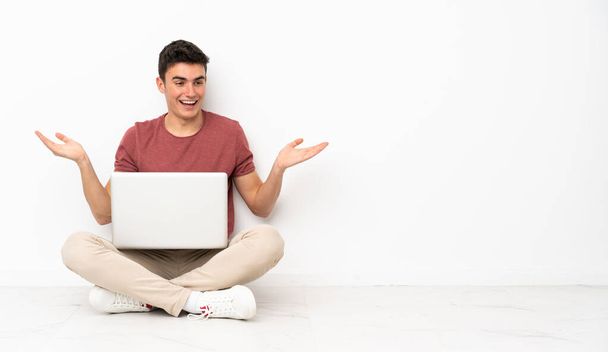 Teenager man sitting on the flor with his laptop holding copyspace with two hands - Foto, Imagem