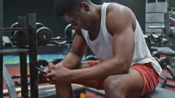 Boční pohled na mladý svalnatý afro muž sedí na lavičce, používá telefon a relaxuje po cvičení - Záběry, video