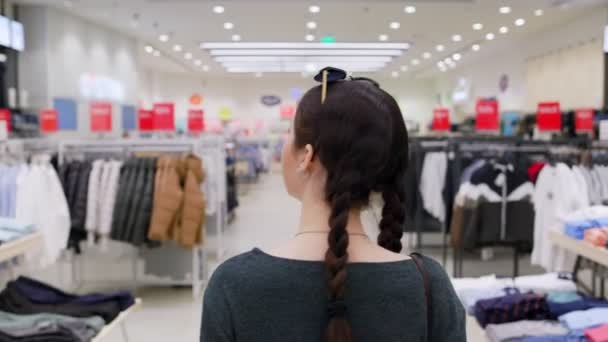 Una joven mujer bonita está caminando por una tienda de ropa y mirando perchas de ropa. En tiempo real. Vista trasera. El concepto de consumismo y compras. - Imágenes, Vídeo