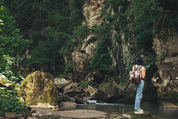 Travel and wanderlust. Stylish woman traveler with backpack standing on background of river in mountains. Young female in casual cloth and hat relaxing on rocks at river, back view. Exploring woods - Photo, Image