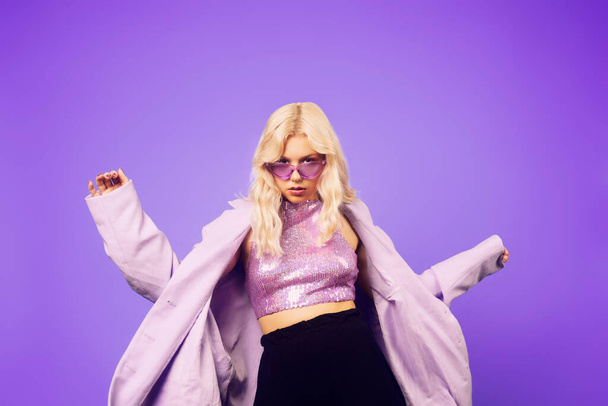 Дівчина в фіолетовому піджаку на фіолетовому тлі в 70-х, 80-х, 90-х роках, стилі макіяж, довгі срібні сережки та фіолетові сонцезахисні окуляри та рожевий піджак. Жінка танцює, вечірка, музика
. - Фото, зображення