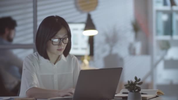 Asyalı iş kadınının karanlık ofisinde masasında oturup dizüstü bilgisayarla çalışırken, sonra gözlüğünü çıkarıp, gözlerini ovalarken görüntüsü. - Video, Çekim