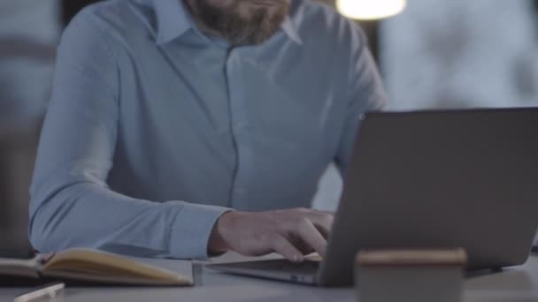 Отслеживание снимка уставшего офисного работника с бородой, сидящего за столом и работающего над ноутбуком вечером, затем снимающего очки и трящего глаза - Кадры, видео
