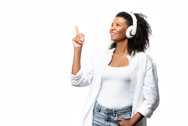 Χαμογελώντας αφροαμερικανή γυναίκα με ακουστικά δείχνοντας με το δάχτυλο απομονώνονται σε λευκό  - Φωτογραφία, εικόνα