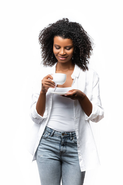 Mujer afroamericana sonriente mirando una taza de café aislada en blanco  - Foto, imagen