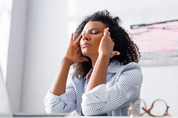 Mujer afroamericana cansada que sufre de dolor de cabeza cerca de la computadora portátil y anteojos en primer plano borrosa  - Foto, imagen