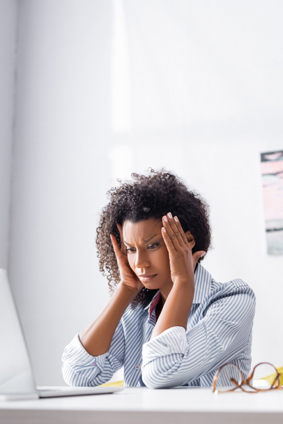 Afro-américain freelance sentiment maux de tête près d'un ordinateur portable et des lunettes sur le premier plan flou  - Photo, image