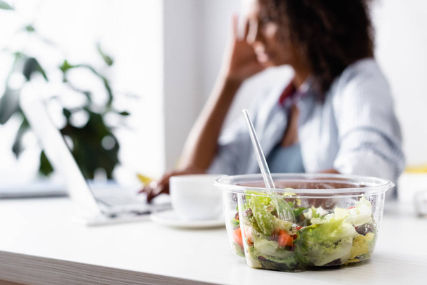 Takeaway salada perto de Africano americano freelancer usando laptop em fundo borrado  - Foto, Imagem