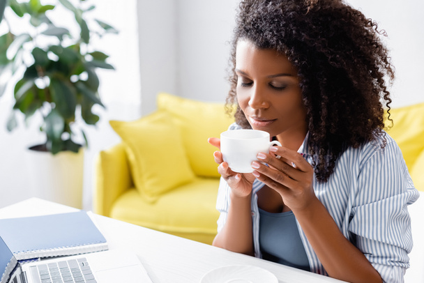 Freelancer afroamericano sosteniendo taza de café cerca de la computadora portátil  - Foto, Imagen