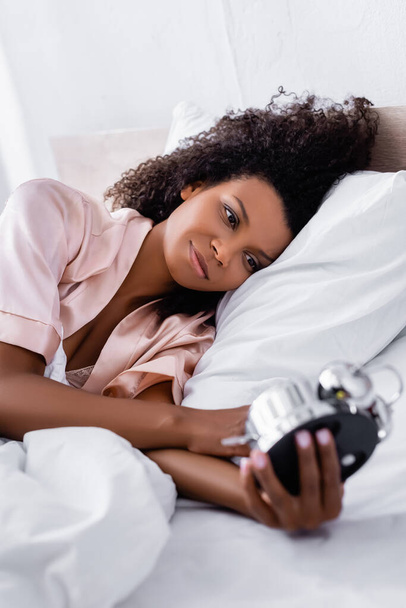 Африканська американка тримає будильник на розмитому передньому плані, коли лежить на ліжку.  - Фото, зображення