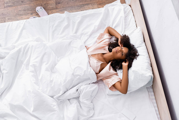 アフリカ系アメリカ人女性のトップビューは、スマートフォンの近くのベッドでストレッチ  - 写真・画像