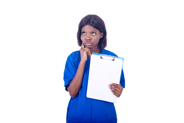 una giovane infermiera affascinante in un camice da laboratorio in piedi su uno sfondo bianco con stetoscopio pensandoci mentre tiene in mano una cartella - Foto, immagini